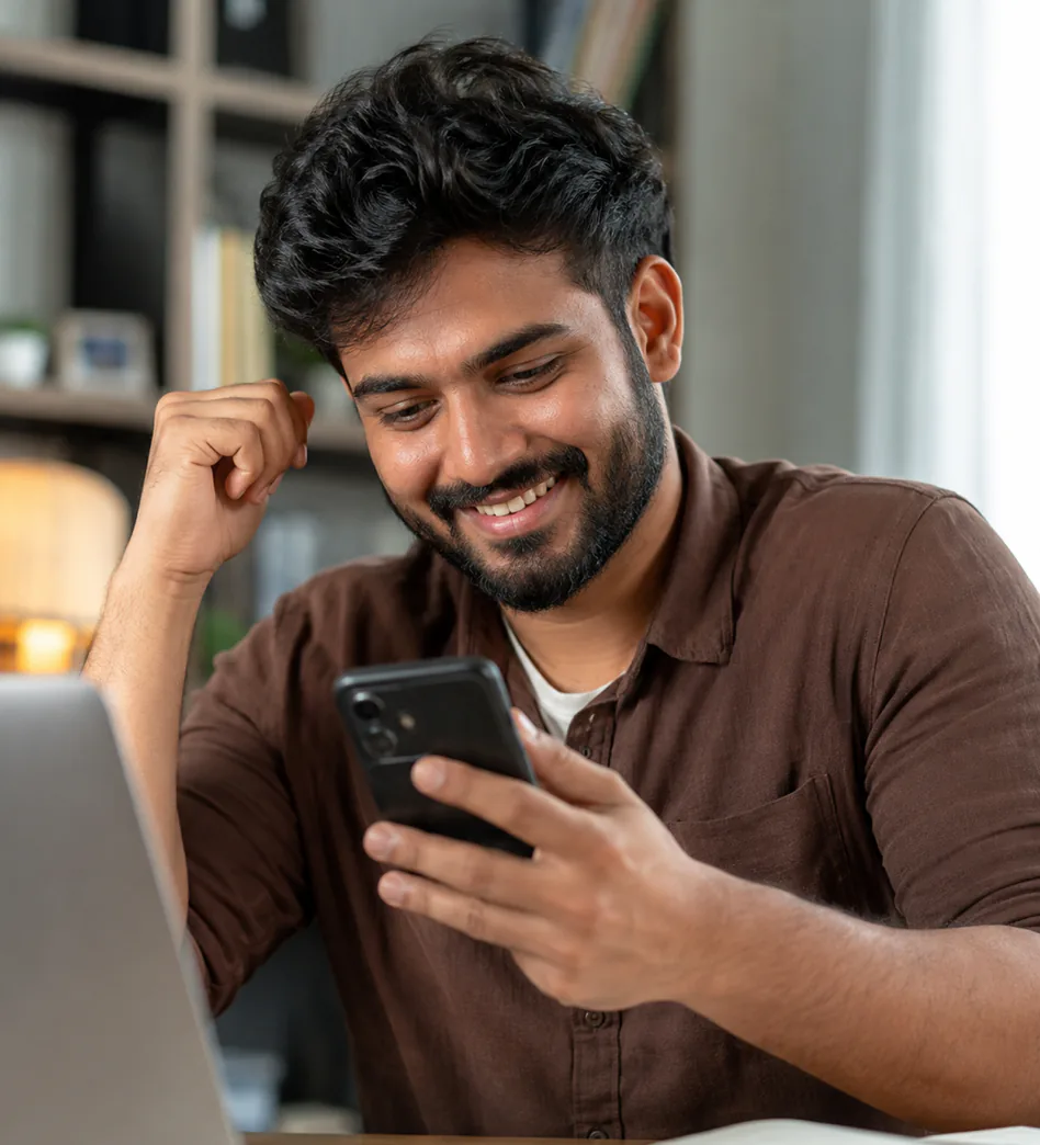 Young man at his desk beside a laptop, smiling at his phone with a fist raised, after reading his career answer from the AstroChat Live AI astrologer